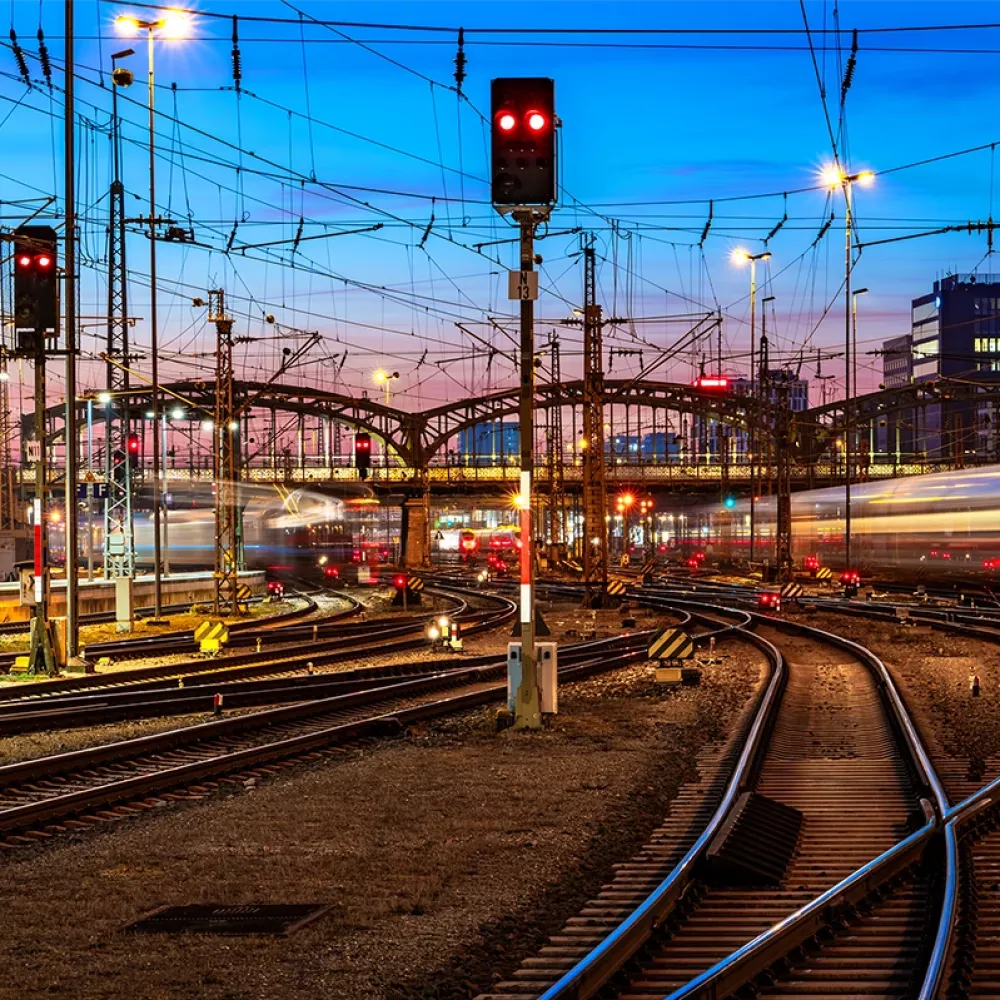 Railway junction at dusk with multiple tracks, overhead power lines, red signal lights, and blurred trains passing through an urban station area.