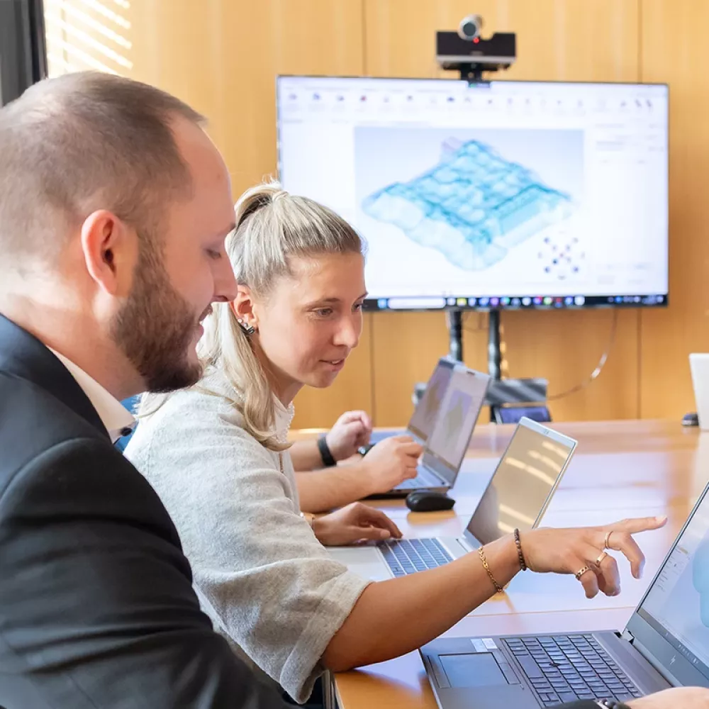 Three people working on laptops in a meeting room while a presenter stands near a large screen displaying a 3D model.