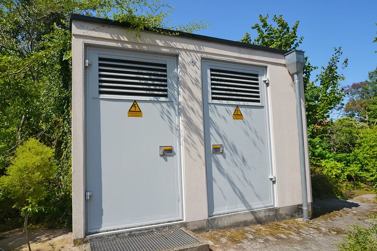 Small gray building with two doors each having ventilation grilles and high voltage warning signs, surrounded by trees.